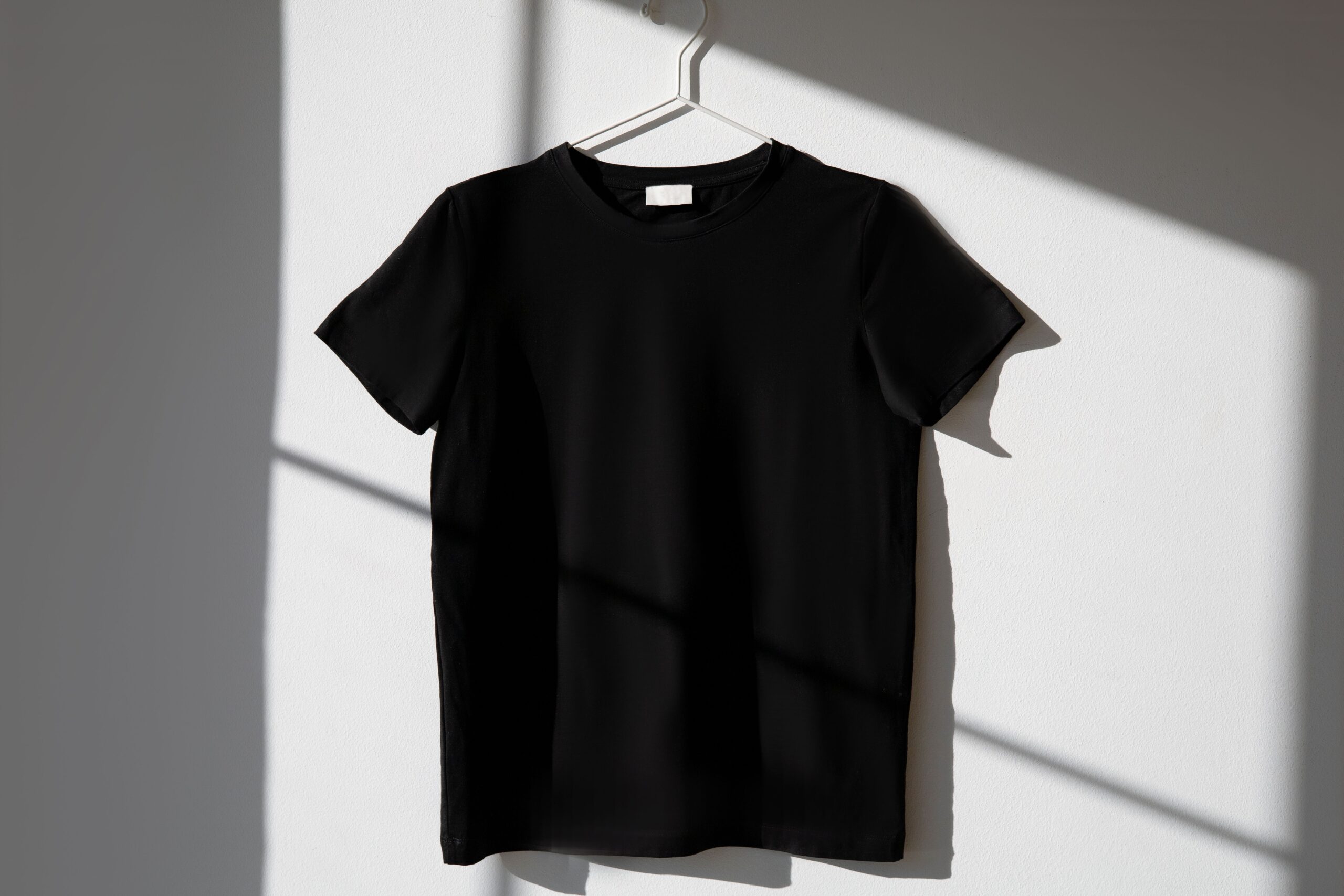 Home Minimalist image of a black t-shirt on a hanger with dramatic shadows on a white wall.